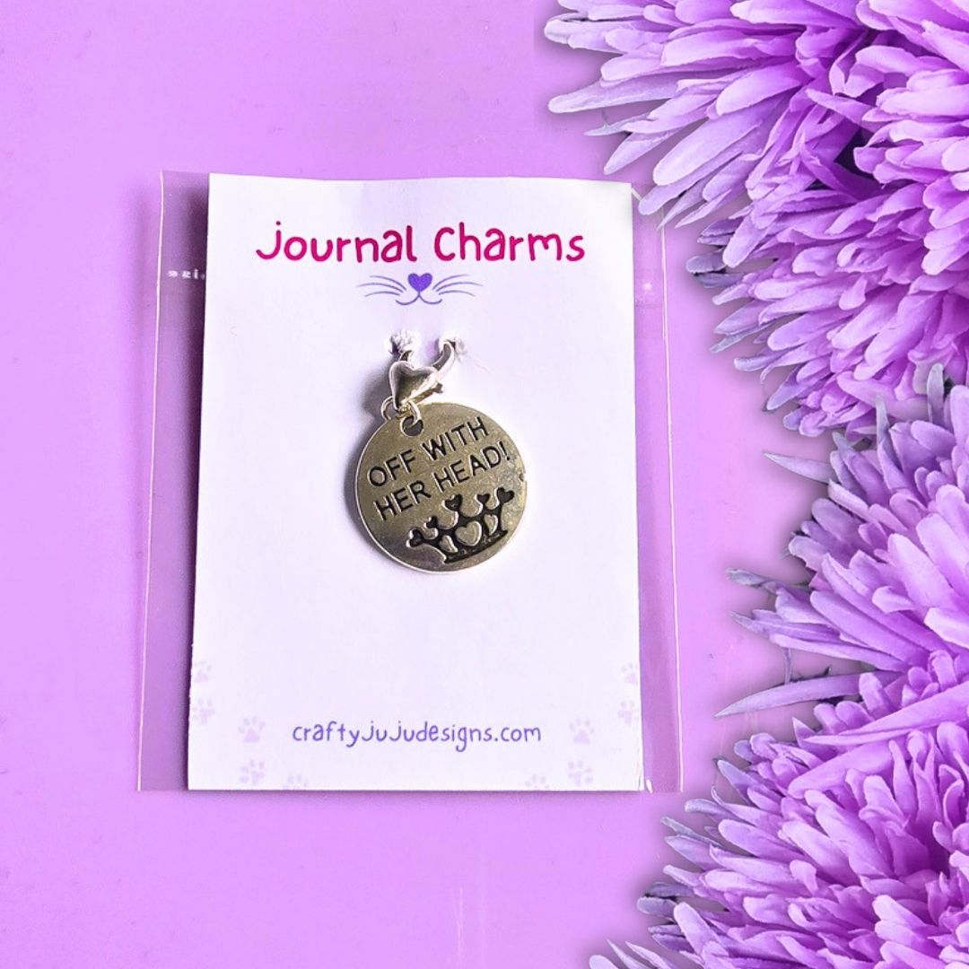 A round silver-toned metal journal charm with the engraved text 'OFF WITH HER HEAD!', displayed on a white card labeled 'Journal Charms' by craftyJudeDesigns.com, set against a purple background with purple flowers.