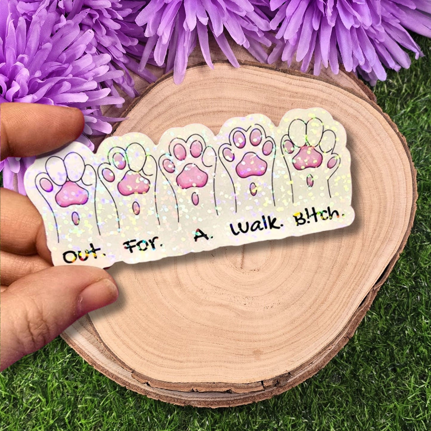 Sticker with paw prints and text held by a hand on a wooden surface with purple flowers in the background.