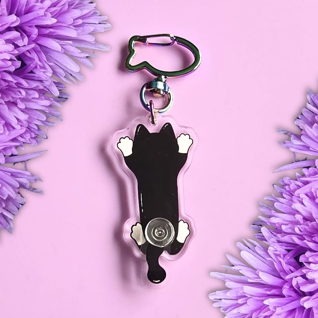 back view of a tuxedo cat acrylic keyring with a moveable tail on the top of a purple background with purple flowers