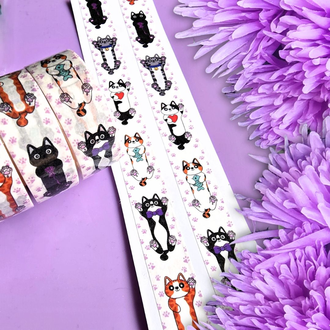 Rolls of decorative washi tape with cute cartoon cat illustrations is displayed on a lavender background next to purple flowers. The tape features black, orange, and white cats with purple and teal accessories, alongside small pink paw prints.