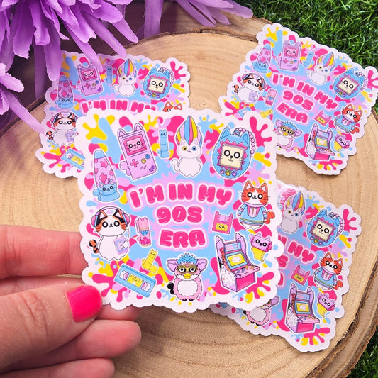 A hand with pink-painted nails and a paw print tattoo is holding a colorful sticker in front of a wooden slab resting on green artificial grass. The sticker has a playful 90s theme, featuring cute cartoon cats and nostalgic icons like a Tamagotchi, video tape, arcade machine, and a Furby-style creature. In the center, bold pink text reads: “I’M IN MY 90s ERA.” The background is bright and fun, with splashes of neon yellow, pink, & blue. In the upper left, a bunch of soft purple flowers adds a vibrant touch