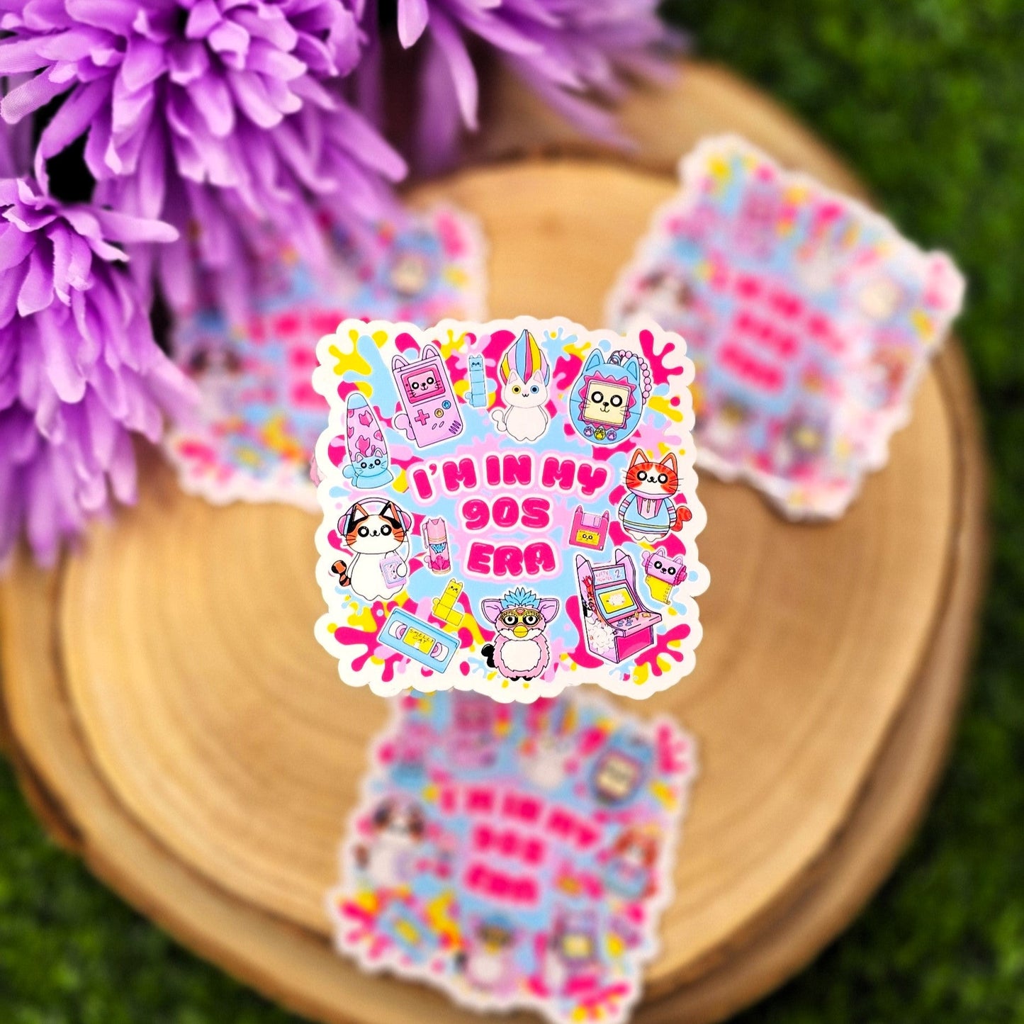 A colorful, die-cut sticker is displayed on a round wooden slab, which rests on vibrant green grass. Behind the wood slab are vivid purple flowers with soft petals. The sticker features a playful, 1990s theme with cute cat characters and nostalgic icons such as a Tamagotchi, video tape, arcade machine, and a character resembling a Furby. In bold pink text in the center, it reads: "I'M IN MY 90s ERA." The background of the sticker has splashes in yellow, blue, & purple adding to the retro aesthetic.