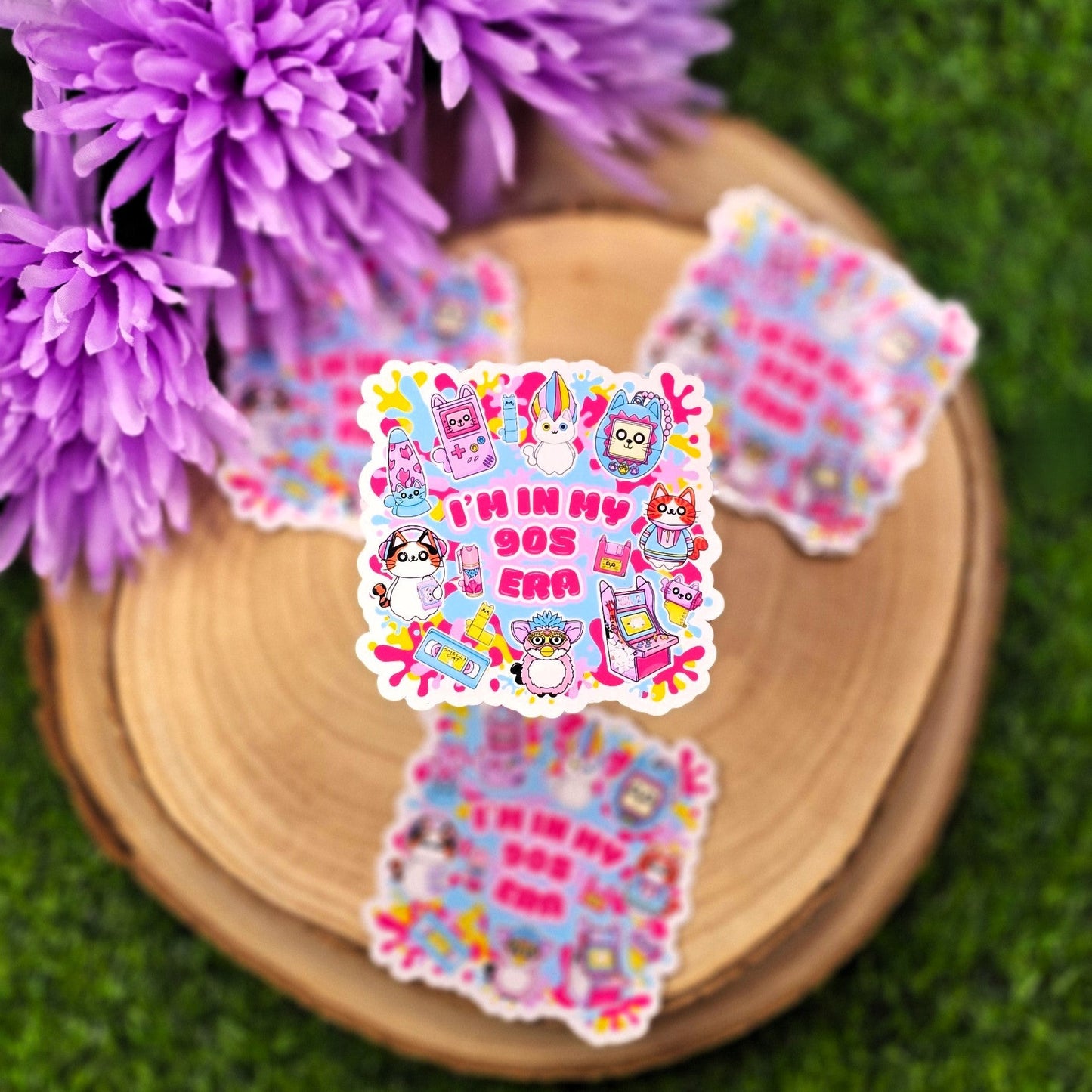 A colorful, die-cut sticker is displayed on a round wooden slab, which rests on vibrant green grass. Behind the wood slab are vivid purple flowers with soft petals. The sticker features a playful, 1990s theme with cute cat characters and nostalgic icons such as a Tamagotchi, video tape, arcade machine, and a character resembling a Furby. In bold pink text in the center, it reads: "I'M IN MY 90s ERA." The background of the sticker has splashes in yellow, blue, & purple adding to the retro aesthetic.