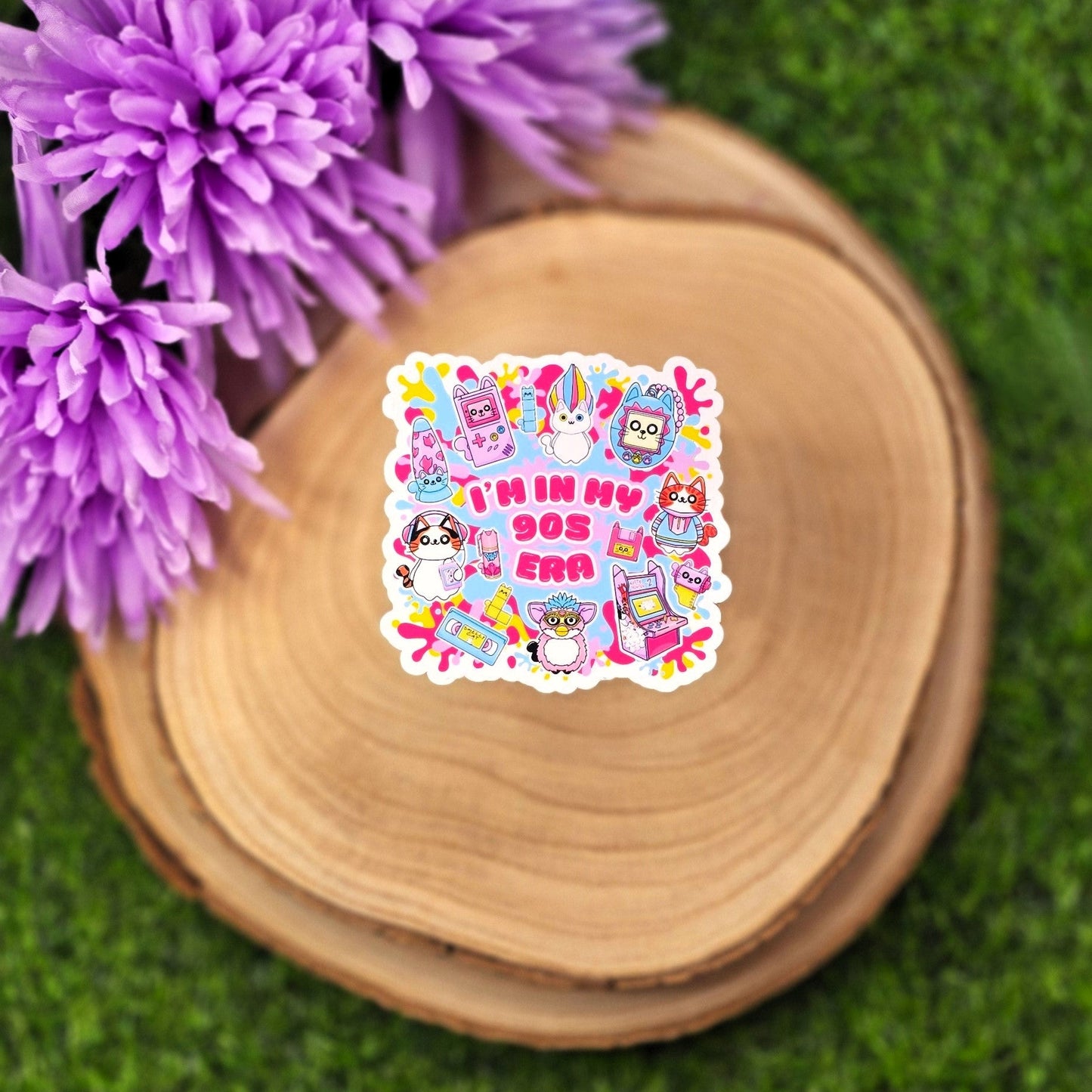 A colorful, die-cut sticker is displayed on a round wooden slab, which rests on vibrant green grass. Behind the wood slab are vivid purple flowers with soft petals. The sticker features a playful, 1990s theme with cute cat characters and nostalgic icons such as a Tamagotchi, video tape, arcade machine, and a character resembling a Furby. In bold pink text in the center, it reads: "I'M IN MY 90s ERA." The background of the sticker has splashes in yellow, blue, & purple adding to the retro aesthetic.