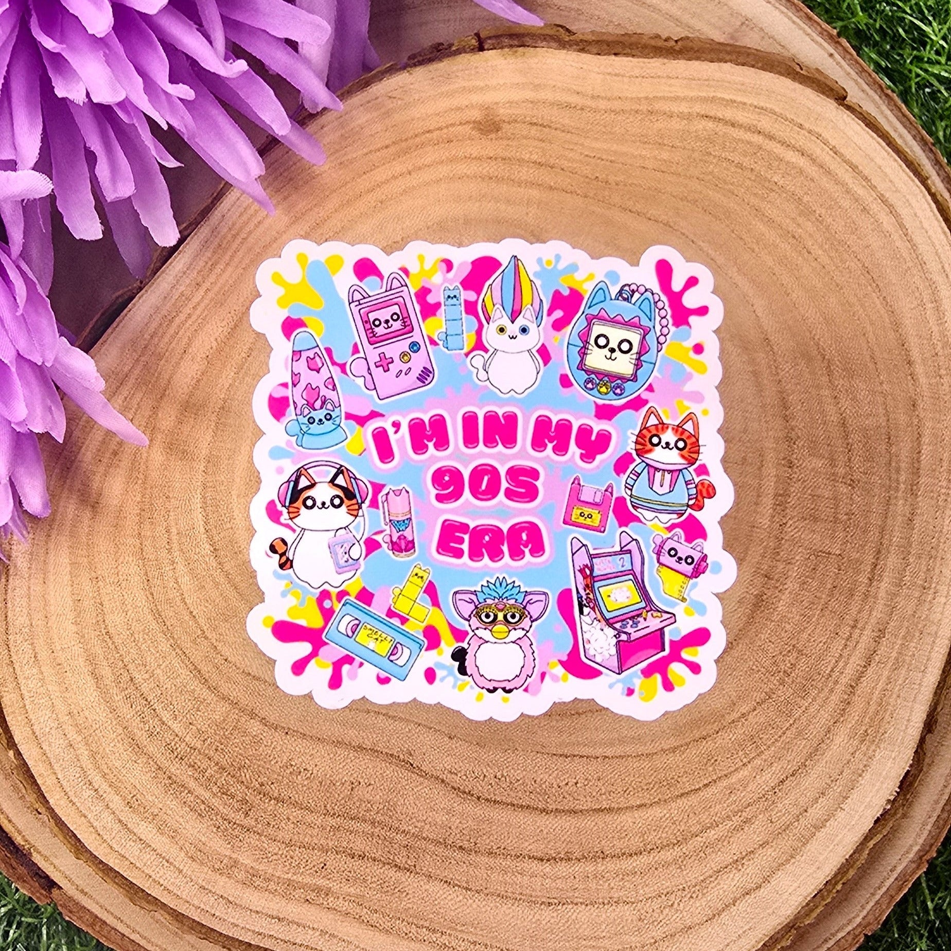 A colorful, die-cut sticker is displayed on a round wooden slab, which rests on vibrant green grass. Behind the wood slab are vivid purple flowers with soft petals. The sticker features a playful, 1990s theme with cute cat characters and nostalgic icons such as a Tamagotchi, video tape, arcade machine, and a character resembling a Furby. In bold pink text in the center, it reads: "I'M IN MY 90s ERA." The background of the sticker has splashes in yellow, blue, & purple adding to the retro aesthetic.