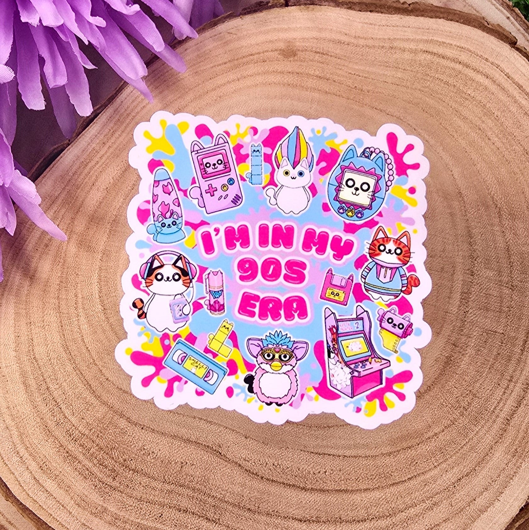 A colorful, die-cut sticker is displayed on a round wooden slab, which rests on vibrant green grass. Behind the wood slab are vivid purple flowers with soft petals. The sticker features a playful, 1990s theme with cute cat characters and nostalgic icons such as a Tamagotchi, video tape, arcade machine, and a character resembling a Furby. In bold pink text in the center, it reads: "I'M IN MY 90s ERA." The background of the sticker has splashes in yellow, blue, & purple adding to the retro aesthetic.
