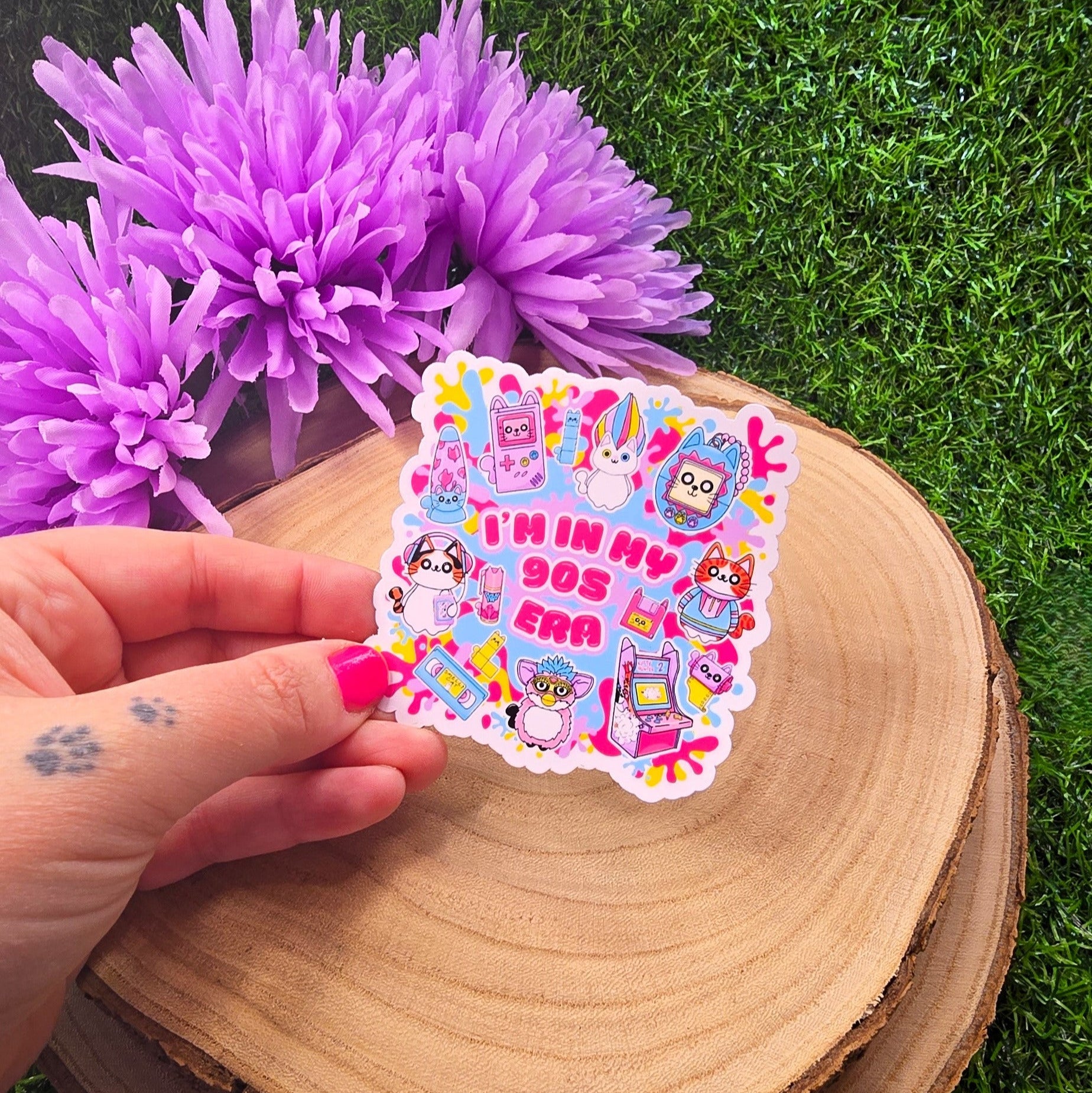 A hand with pink-painted nails and a paw print tattoo is holding a colorful sticker in front of a wooden slab resting on green artificial grass. The sticker has a playful 90s theme, featuring cute cartoon cats and nostalgic icons like a Tamagotchi, video tape, arcade machine, and a Furby-style creature. In the center, bold pink text reads: “I’M IN MY 90s ERA.” The background is bright and fun, with splashes of neon yellow, pink, & blue. In the upper left, a bunch of soft purple flowers adds a vibrant touch