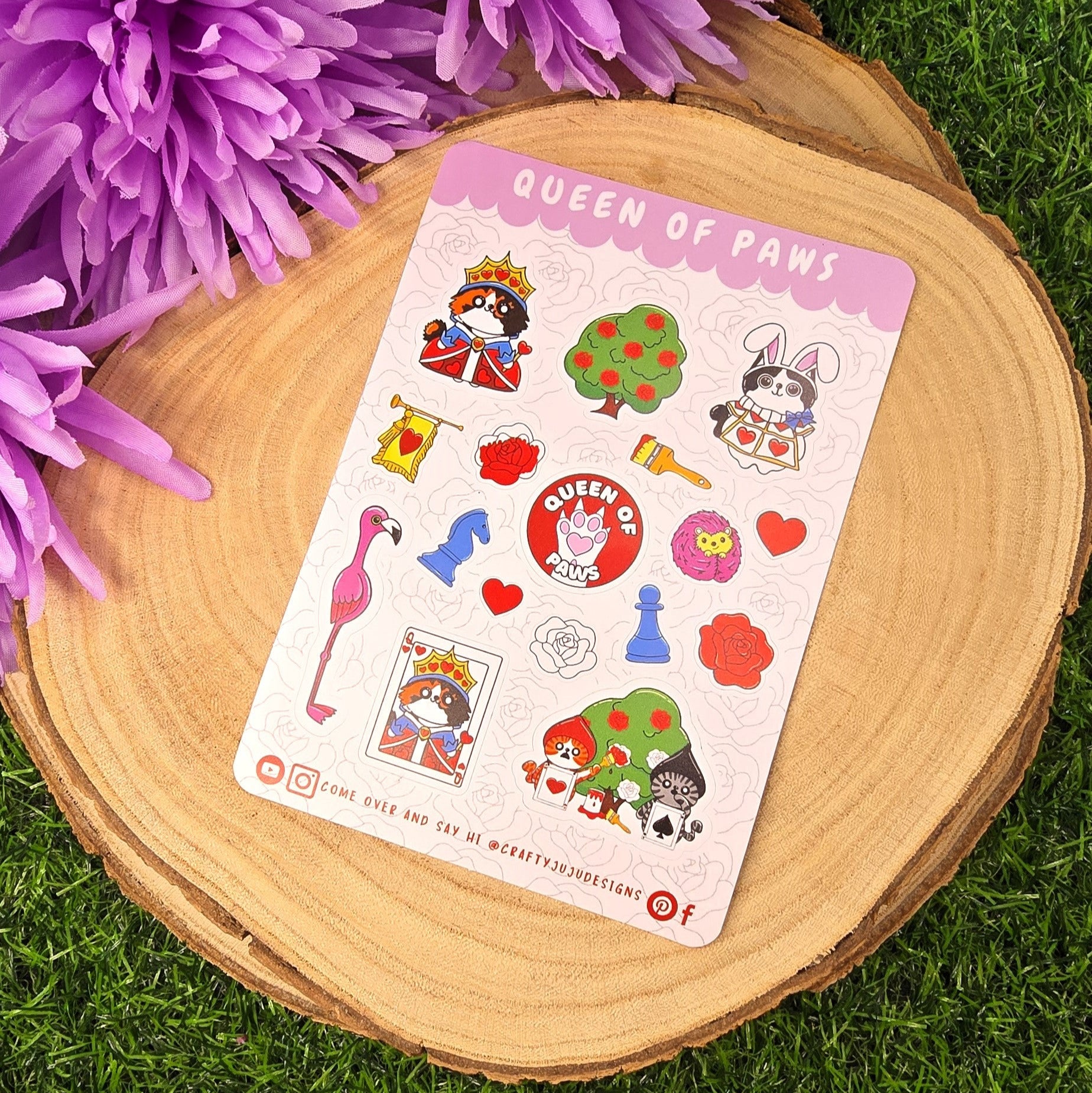 Sticker sheet titled 'Queen of Paws' on a wooden surface with purple flowers layed on a grass green background.