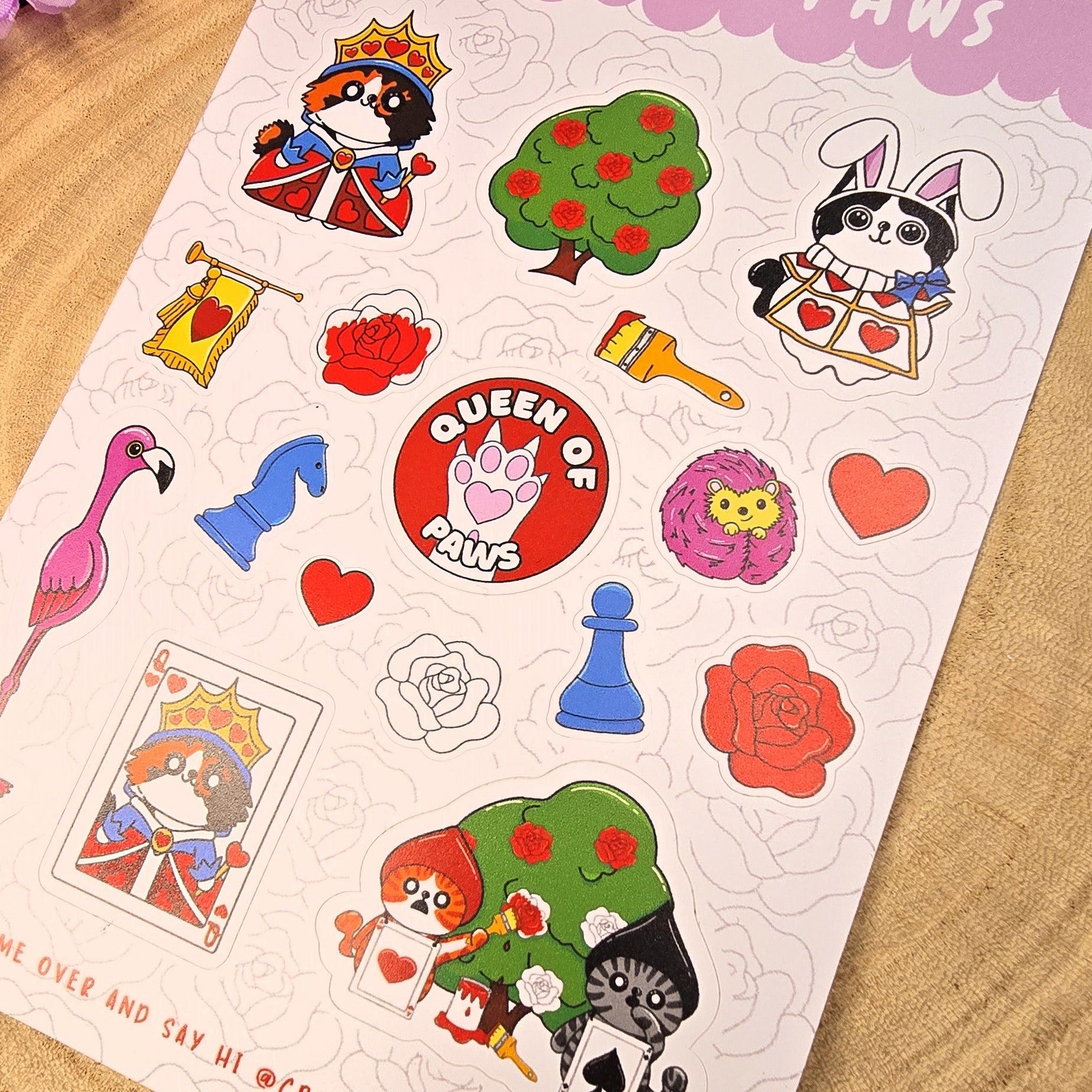 Sticker sheet titled 'Queen of Paws' with various illustrations on a wooden surface with purple flowers in the background.