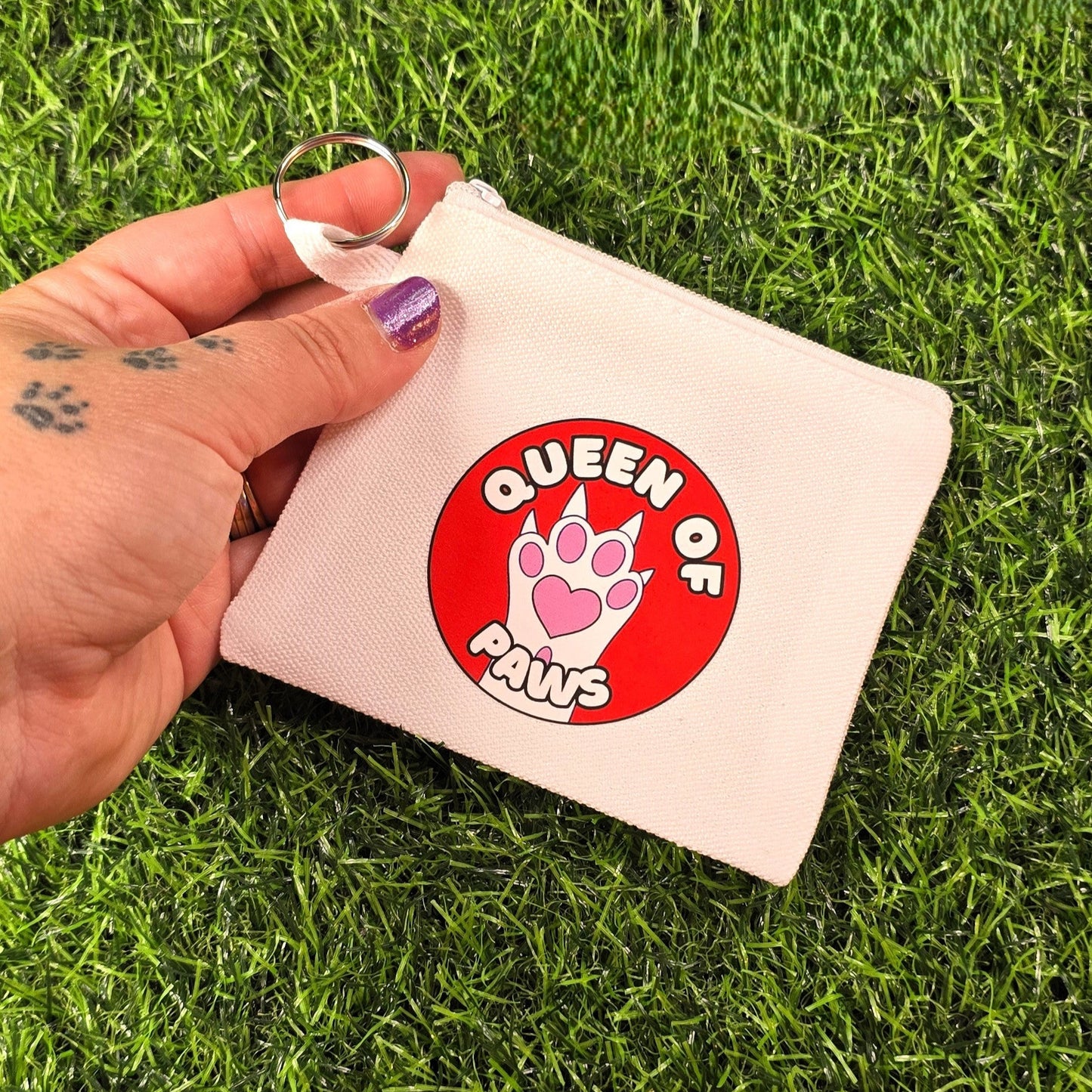 Hand holding a small fabric coin purse with 'Queen of Paws' sticker on over a grass background
