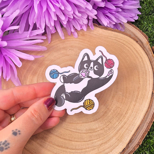 Hand holding a sticker of a cat with yarn balls on a wooden slice with purple flowers in the background.
