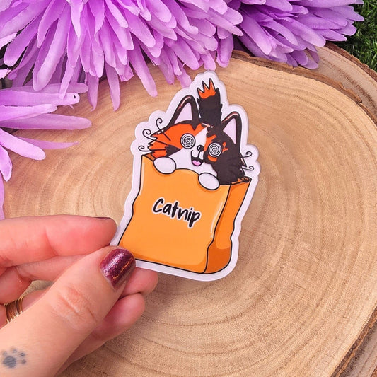 Hand holding a sticker with a cat design labeled 'Catnip' on a wooden surface with purple flowers.