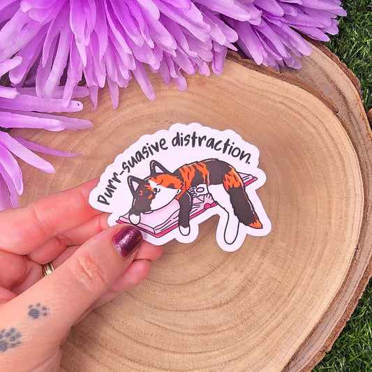 Sticker of a cat on a book held over a wooden surface with purple flowers.