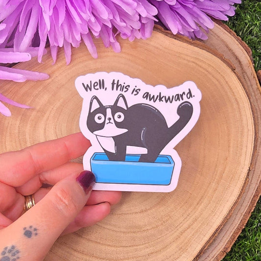 Hand holding a sticker with a cat and text 'Well, this is awkward.' on a wooden surface with purple flowers.