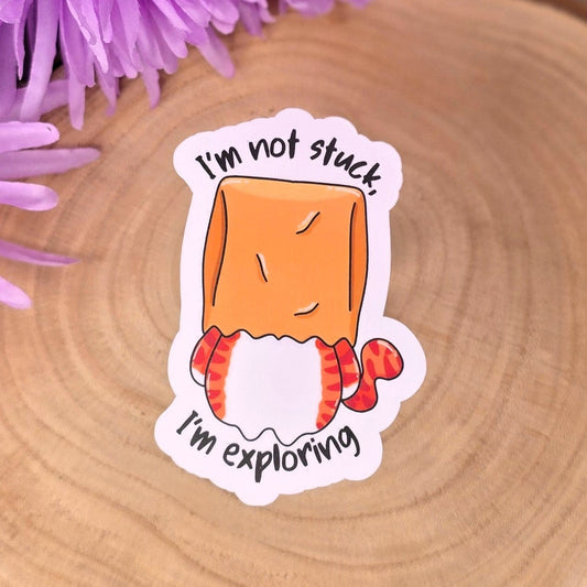 Sticker with a cartoon character and text 'I'm not stuck, I'm exploring' on a wooden surface with purple flowers.