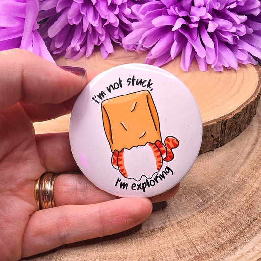 Button badge with cartoon cat character and text 'I'm not stuck, I'm exploring' held by a hand against purple flowers.