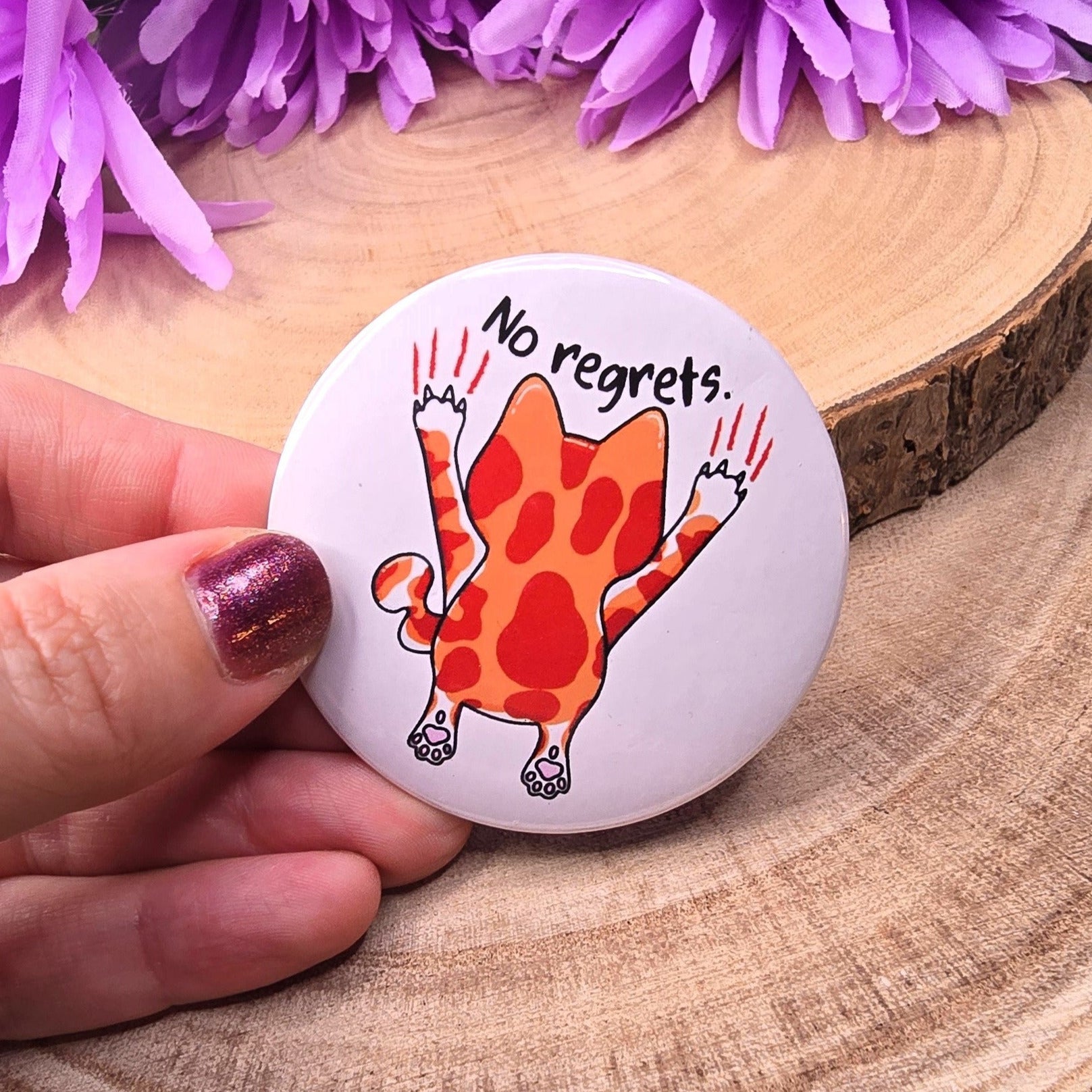 Button badge with a cat illustration and 'No regrets' text on a wooden surface with purple flowers.