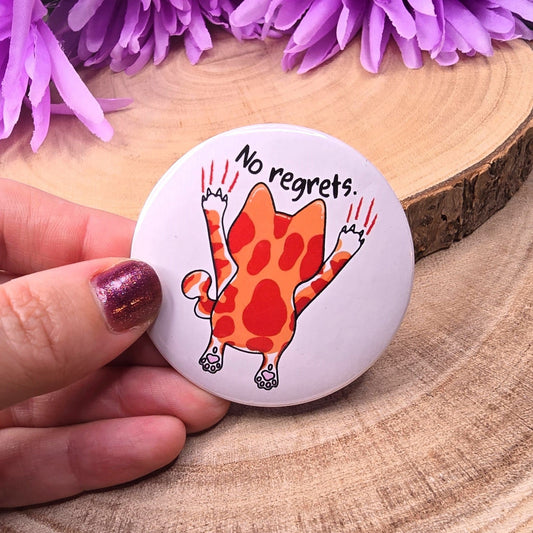 Button badge with a cat illustration and 'No regrets' text on a wooden surface with purple flowers.