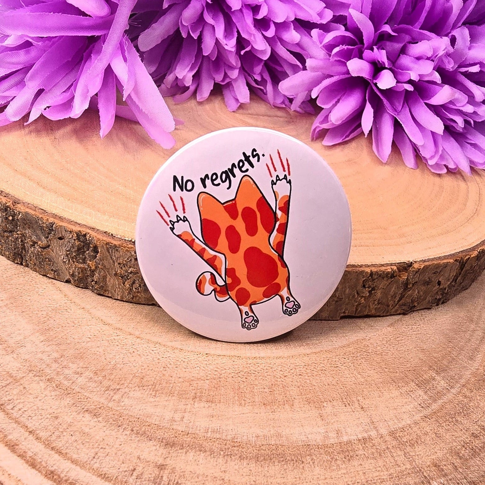 Button badge with a cartoon character and 'No regrets' text on a wooden surface with purple flowers.