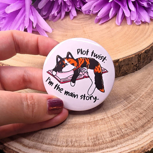 Button with a cat illustration and text held by a hand on a wooden surface with purple flowers.