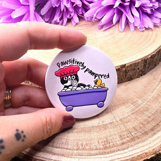 Button badge with cartoon cat in a bath and text 'Pawsitively pampered' held by a hand on a wooden surface with purple flowers in the background.
