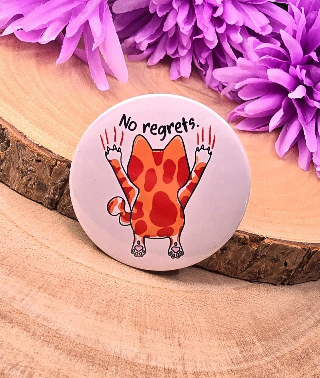 Button with a cat illustration and 'No regrets' text on a wooden surface with purple flowers.