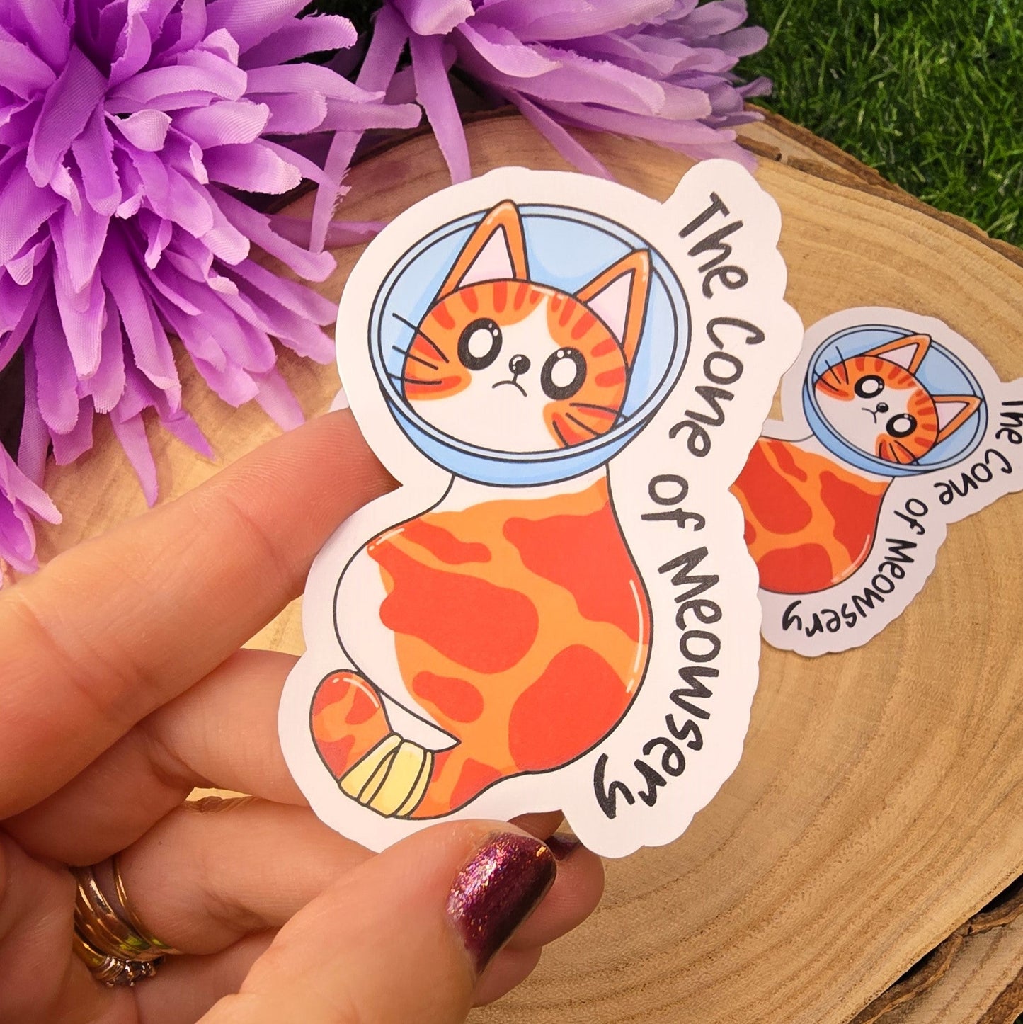 Hand holding a sticker with a cat design and text, surrounded by purple flowers on a wooden surface.