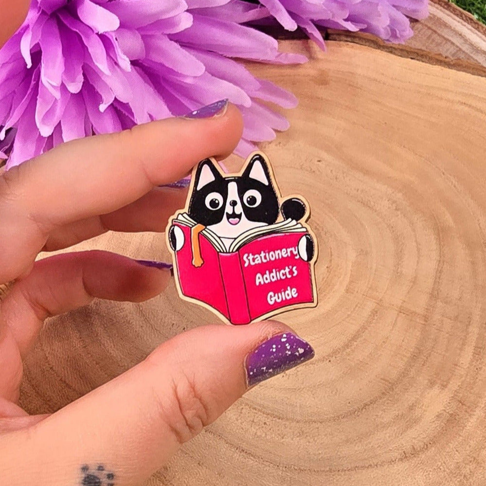 Hand holding a small wooden pin with a black and white cat design, surrounded by purple flowers on a wooden stump.