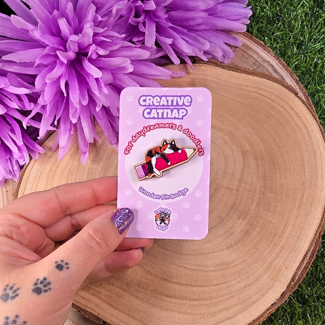Hand holding a 'Creative Catnap' product card with purple flowers in the background 