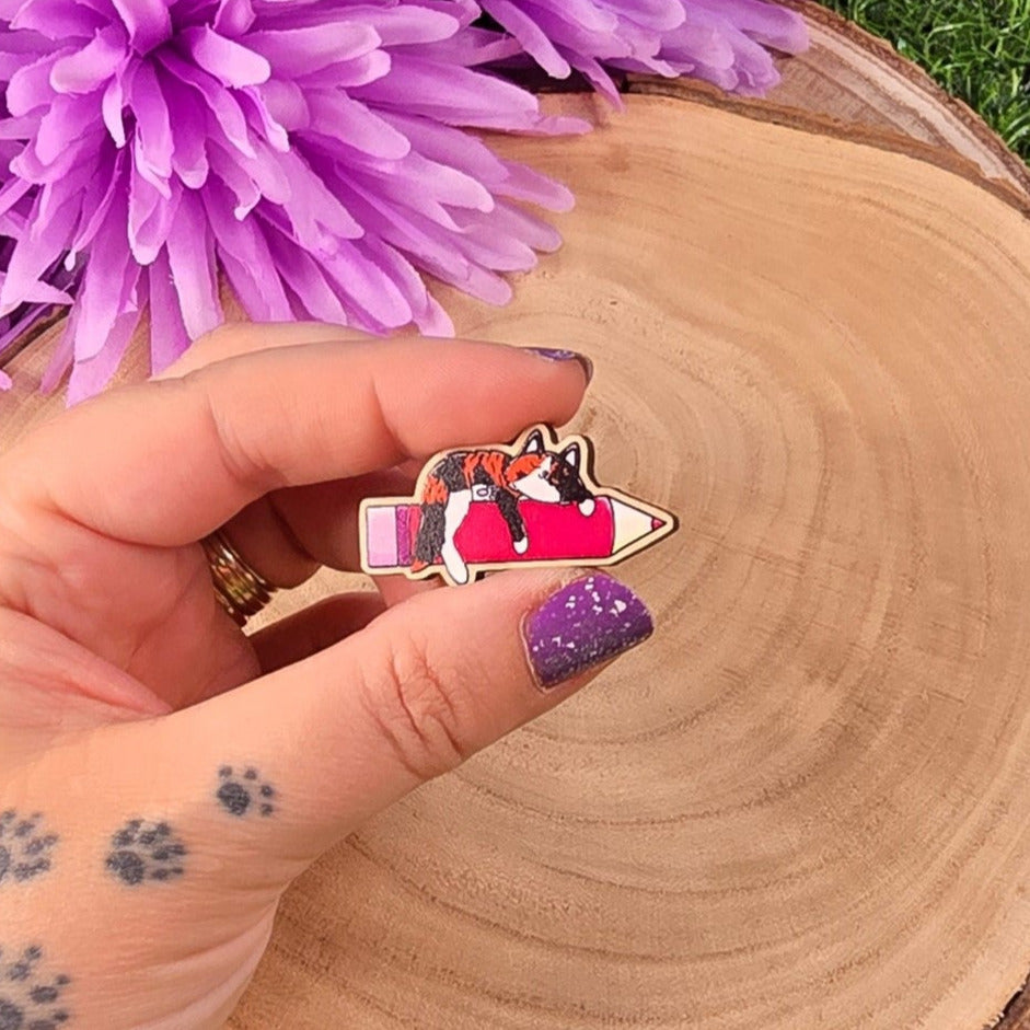 Hand holding a small pink 
pencil with a cat design, surrounded by purple flowers on a grassy background.