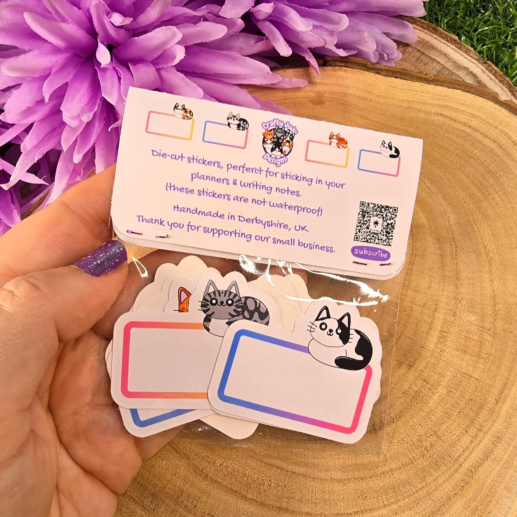 Hand holding a set of die-cut stickers with cat illustrations, placed on a wooden surface with purple flowers in the background.