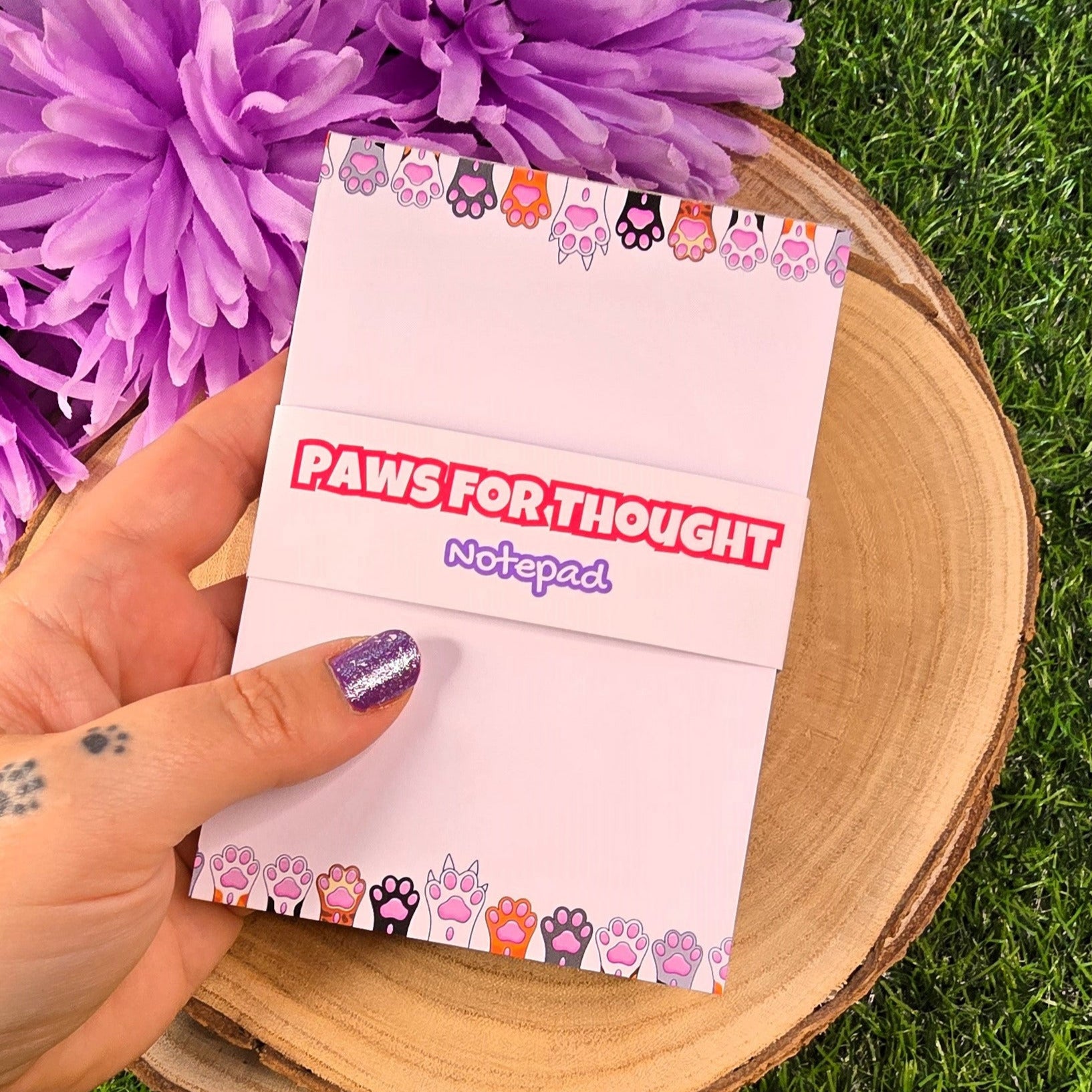 Notepad with 'Paws for Thought' text held by a hand on grass with purple flowers in the background 