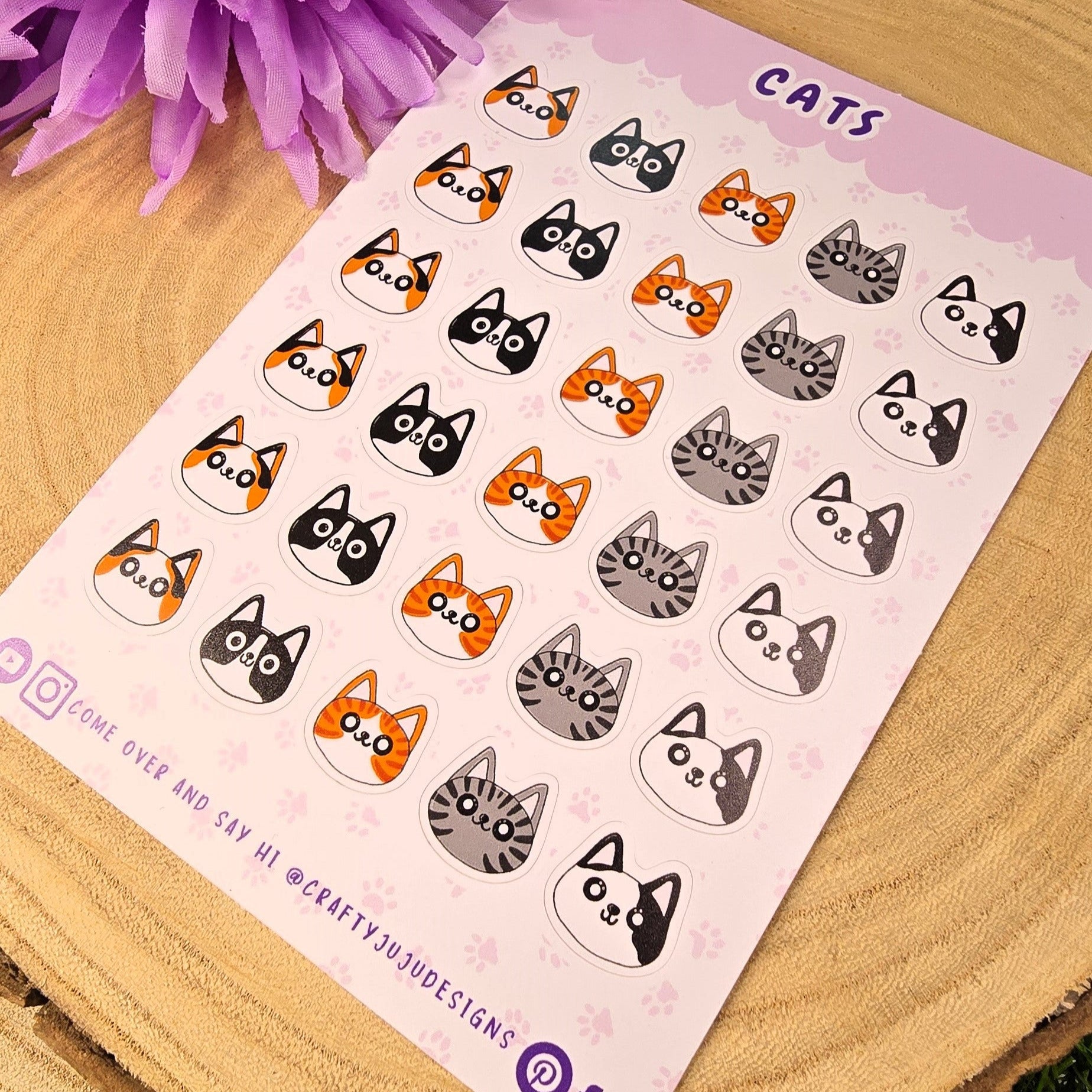 Sheet of cat-themed stickers on a wooden surface with purple flowers in the background