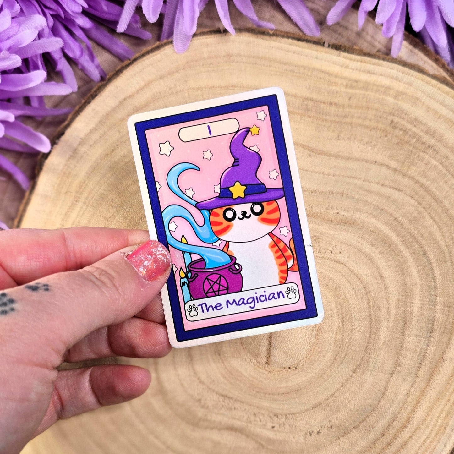 sticker featuring a cat in a wizard hat held by a hand with purple flowers in the background