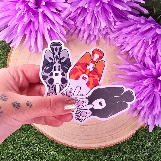 Three cat-shaped stickers on a wooden surface with purple flowers in the background.