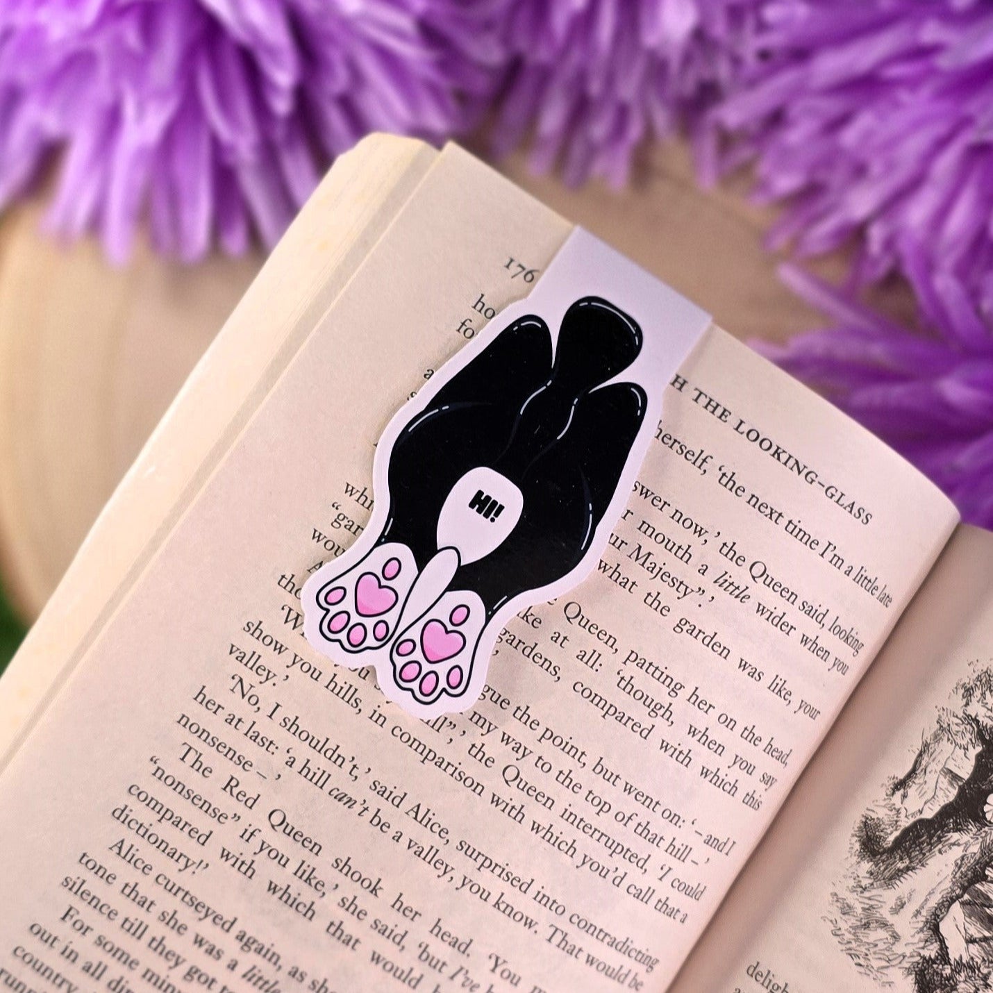 Black and white cat-shaped bookmark with pink paw prints on an open book with purple flowers in the background