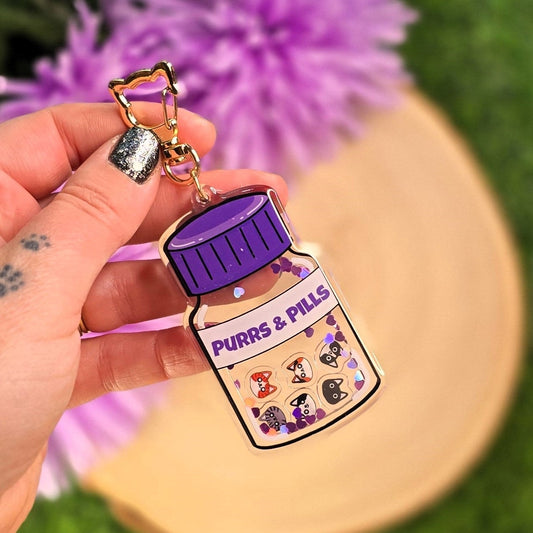 Keychain shaped like a purple pill bottle with cartoon cats on a grassy background