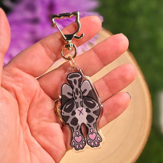 Hand holding a keychain with a cat design against a blurred green and purple background
