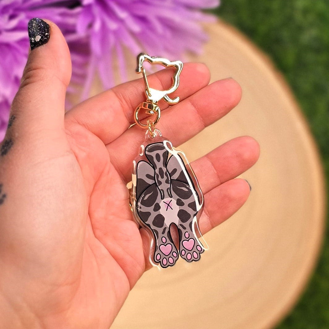 Hand holding a cat-shaped keychain with a star and paw prints, against a blurred background of purple flowers and green grass.
