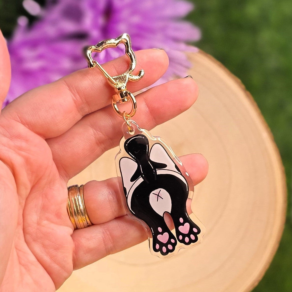 Hand holding a black and white cat-shaped keychain with a blurred purple flower in the background
