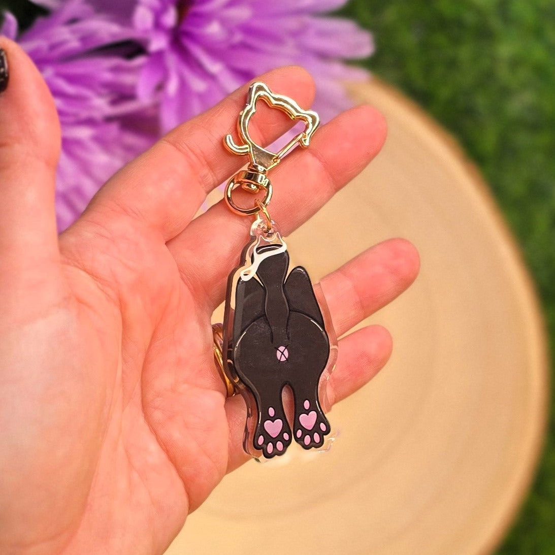 Hand holding a black cat-shaped keychain with pink paw prints against a blurred purple flower and green grass background.