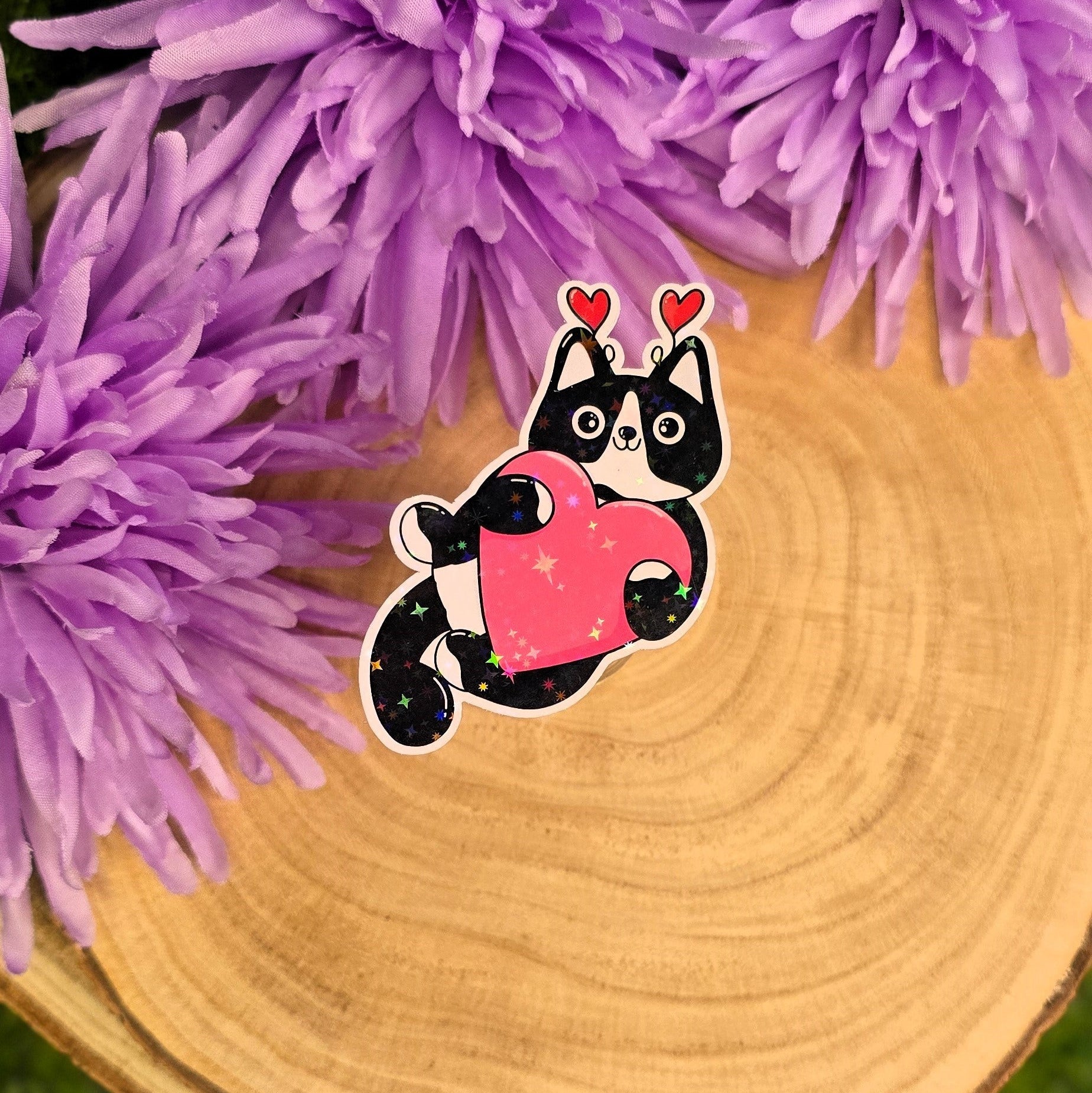 sticker of a black cat with a pink heart on a wooden surface with purple flowers in the background