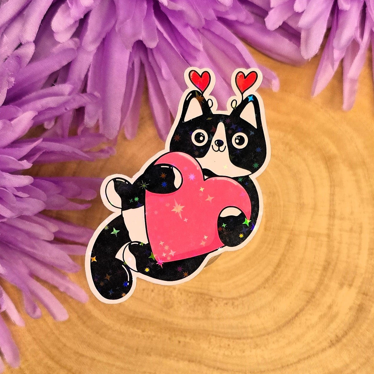 Sticker of a black and white cat holding a pink heart on a wooden surface with purple flowers in the background.