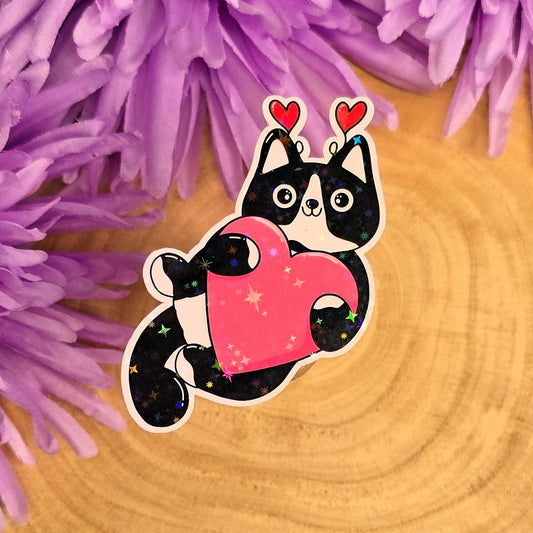 Sticker of a black and white cat holding a pink heart on a wooden surface with purple flowers in the background.