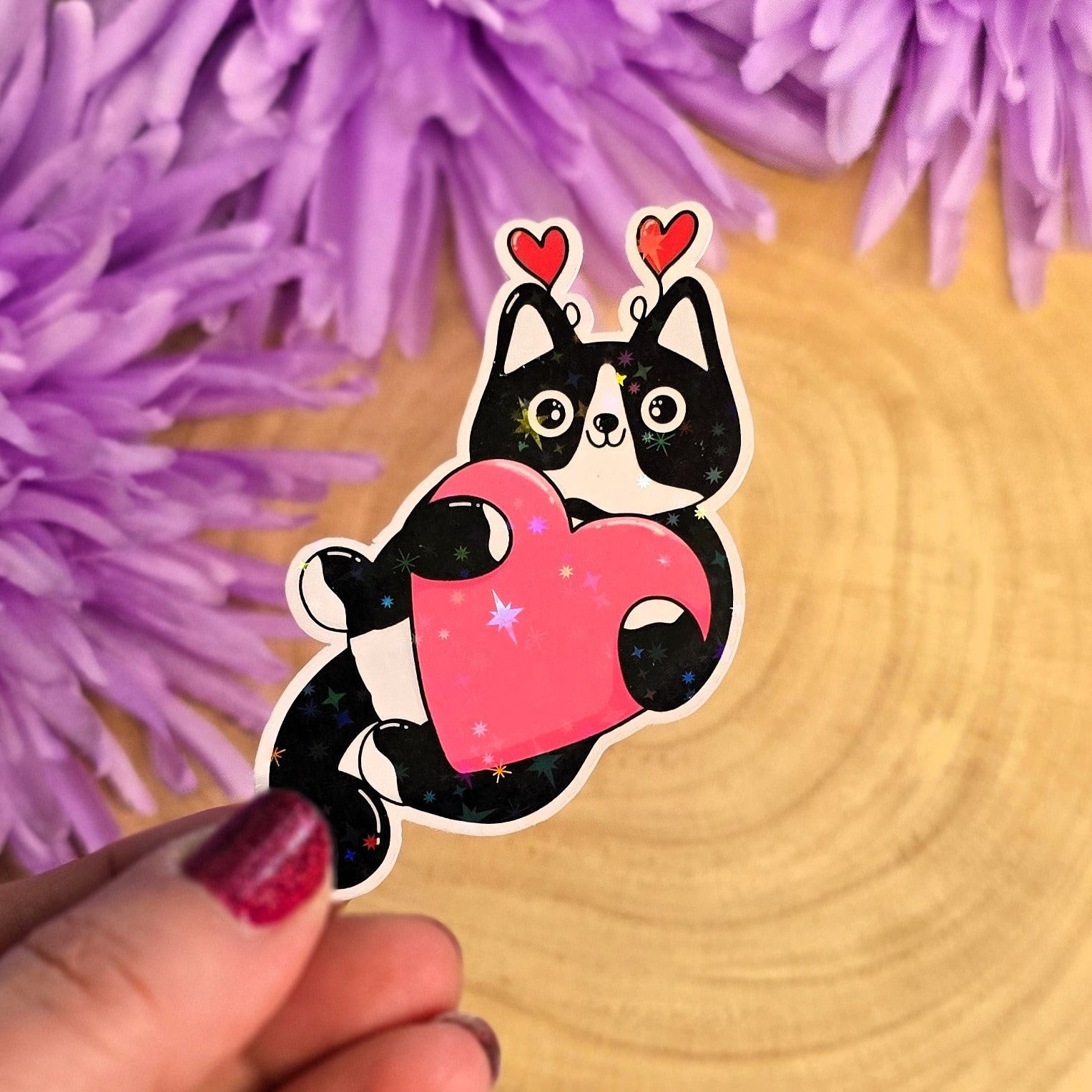 Hand holding a sticker of a cat with a heart, surrounded by purple flowers on a wooden surface.
