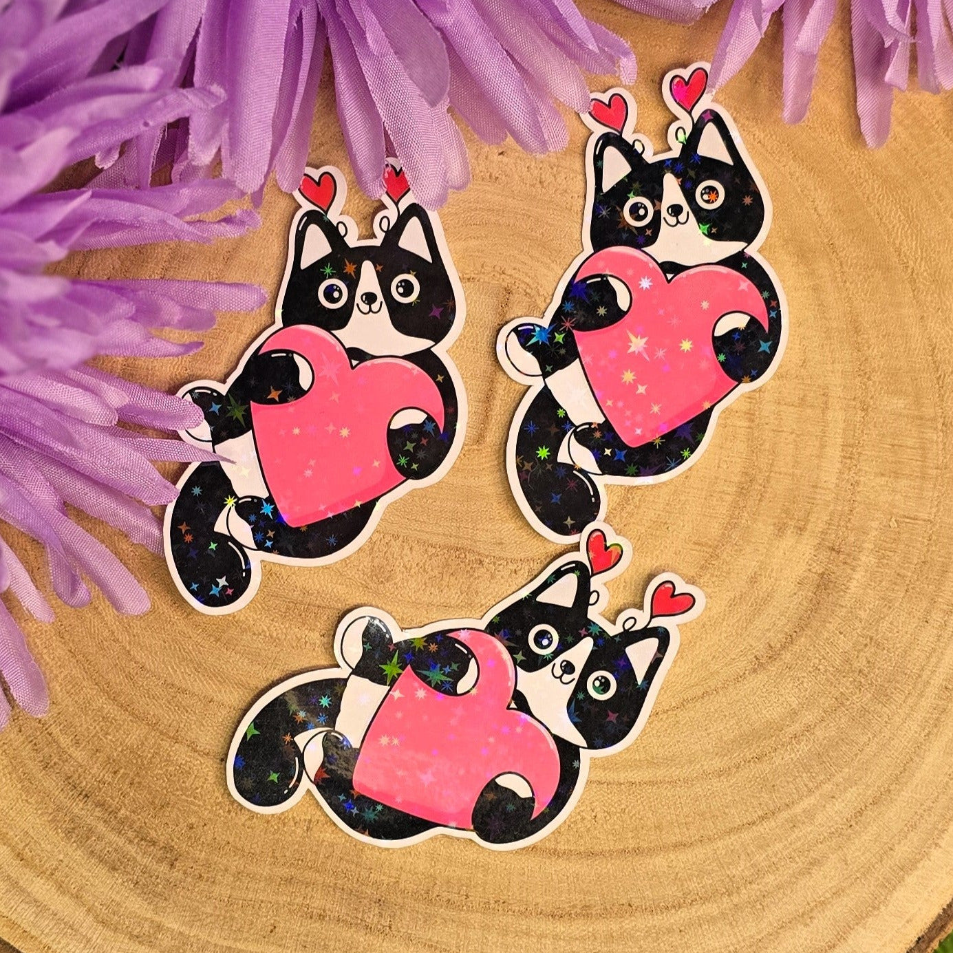 Three cat-shaped stickers with pink hearts on a wooden surface with purple flowers in the background.