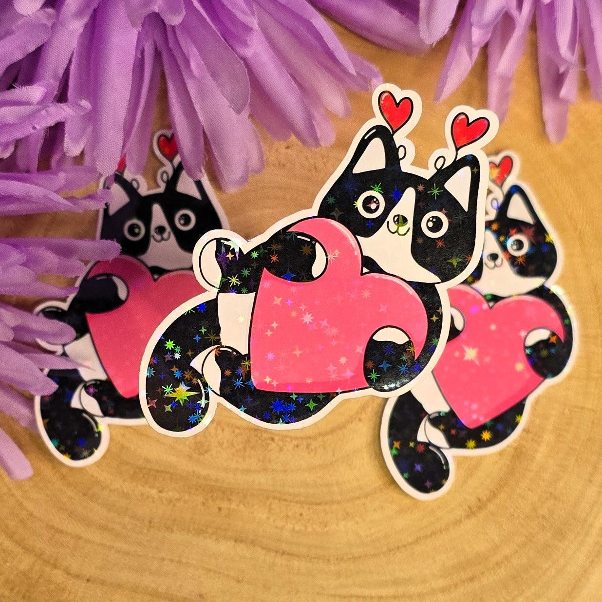 Three black and white cat-shaped stickers with pink hearts on a wooden surface with purple flowers in the background.