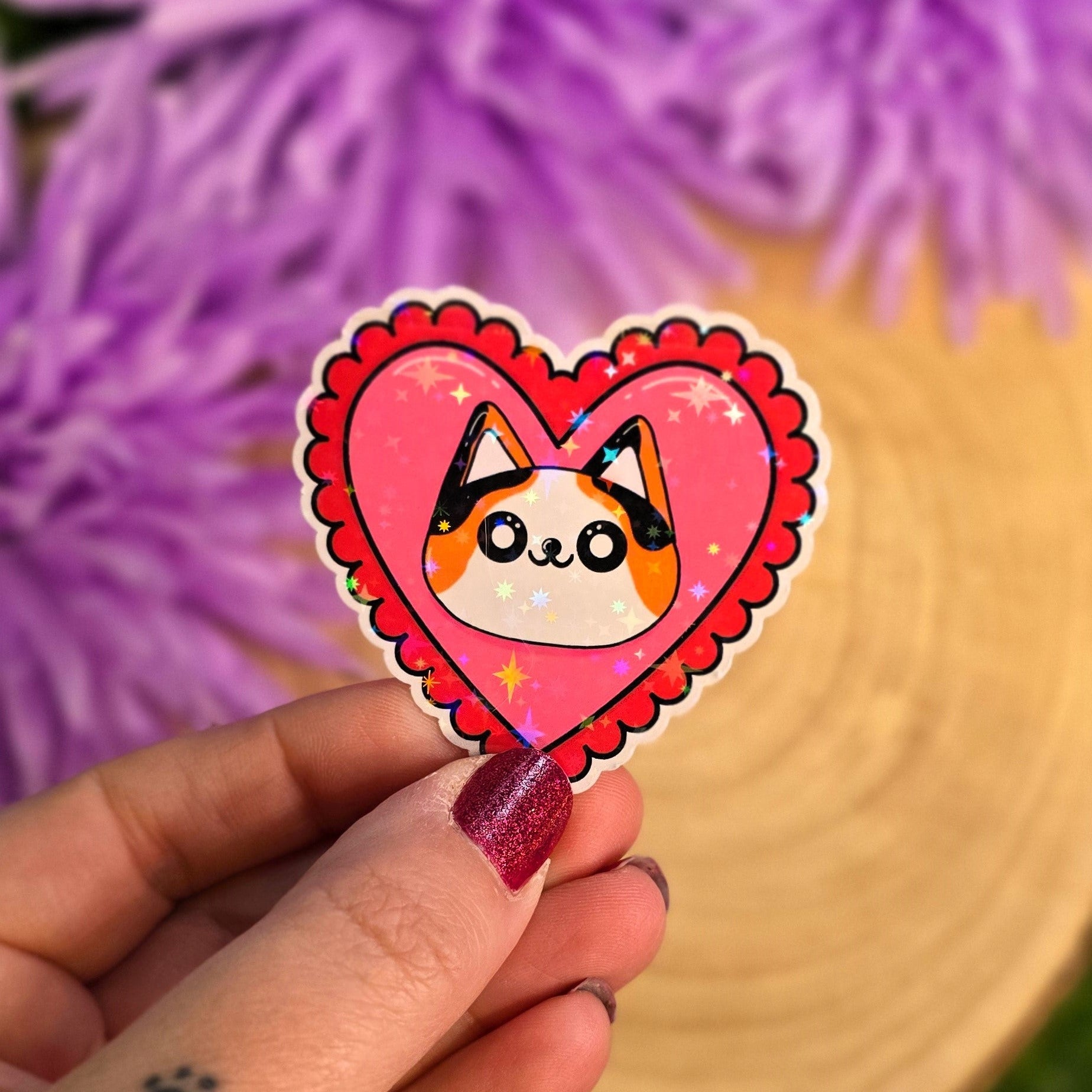 Heart-shaped sticker with a cat design held by a hand against a blurred purple flower background