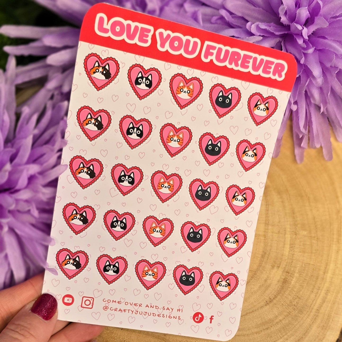 Sheet of heart-shaped stickers with cartoon faces held by a hand in front of purple flowers.