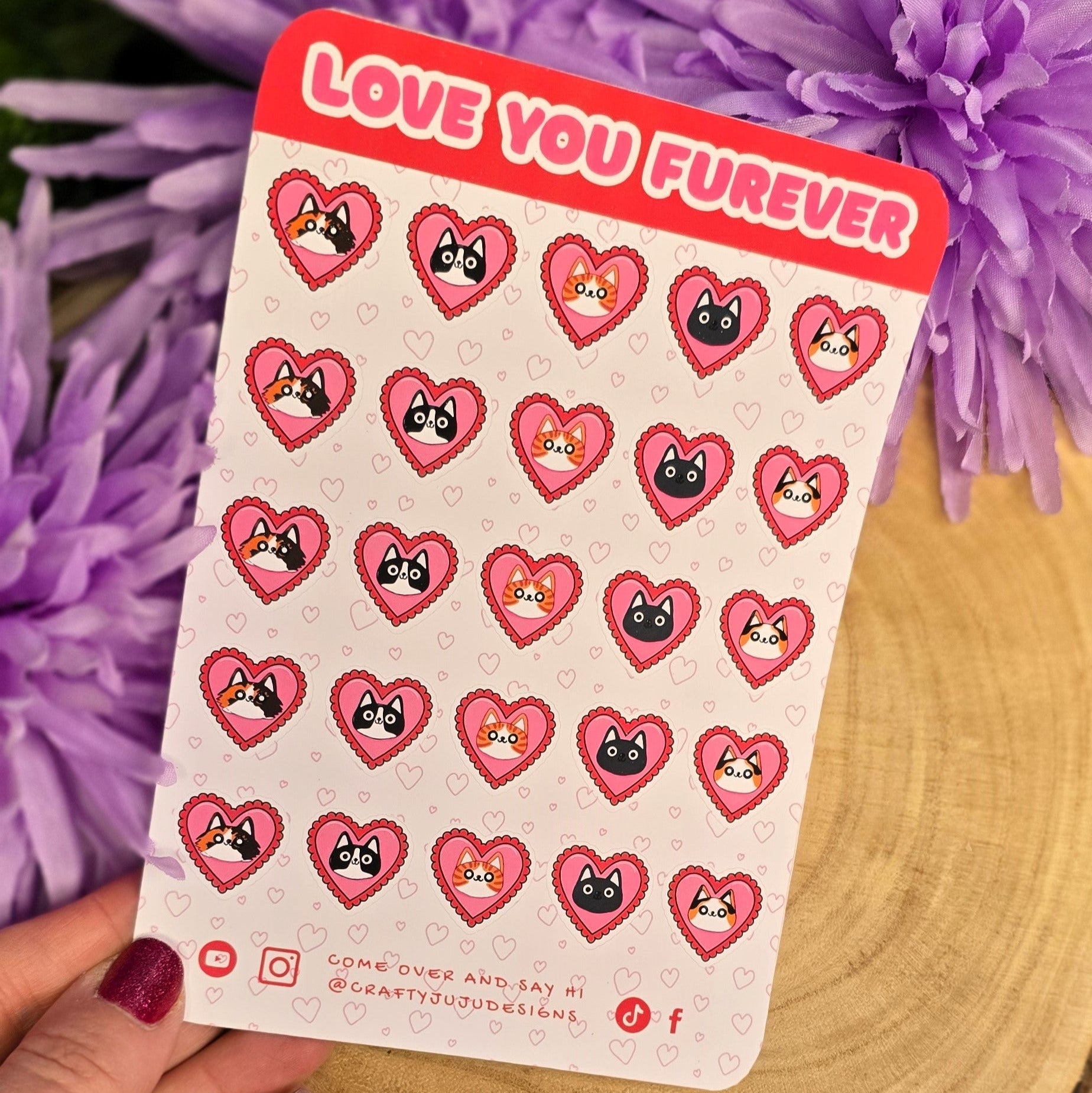 Sheet of heart-shaped stickers with cartoon faces held by a hand in front of purple flowers.